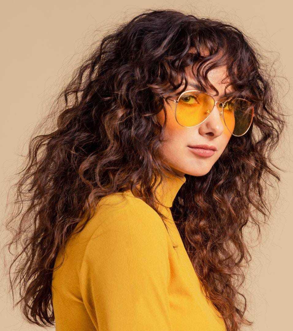 Curly-haired woman in yellow outfit with matching sunglasses, posing confidently.