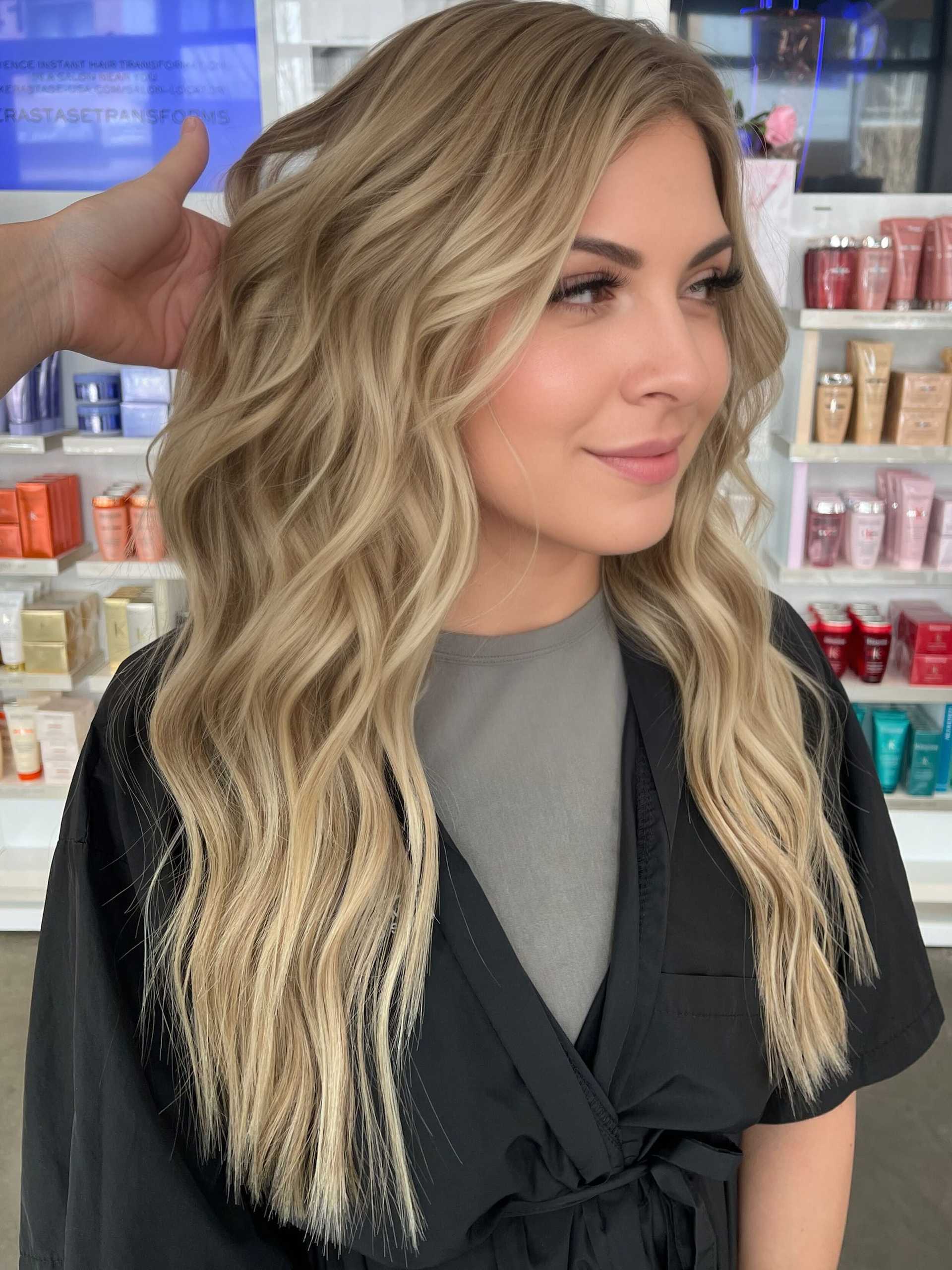 Blonde woman with wavy hair in salon, black cape, surrounded by hair products.