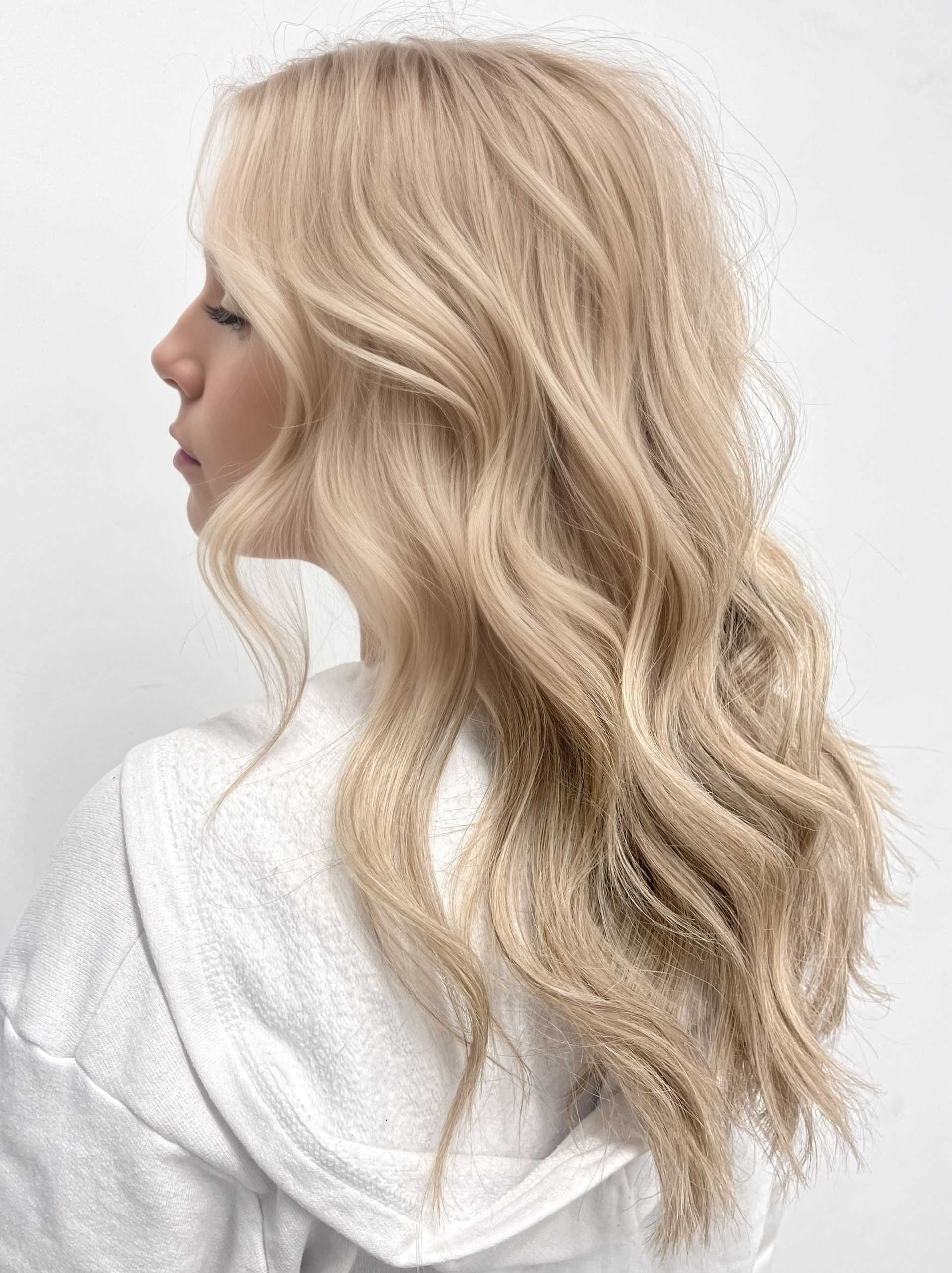 Blonde woman with wavy hair in a white hoodie, viewed from the side against a plain background.