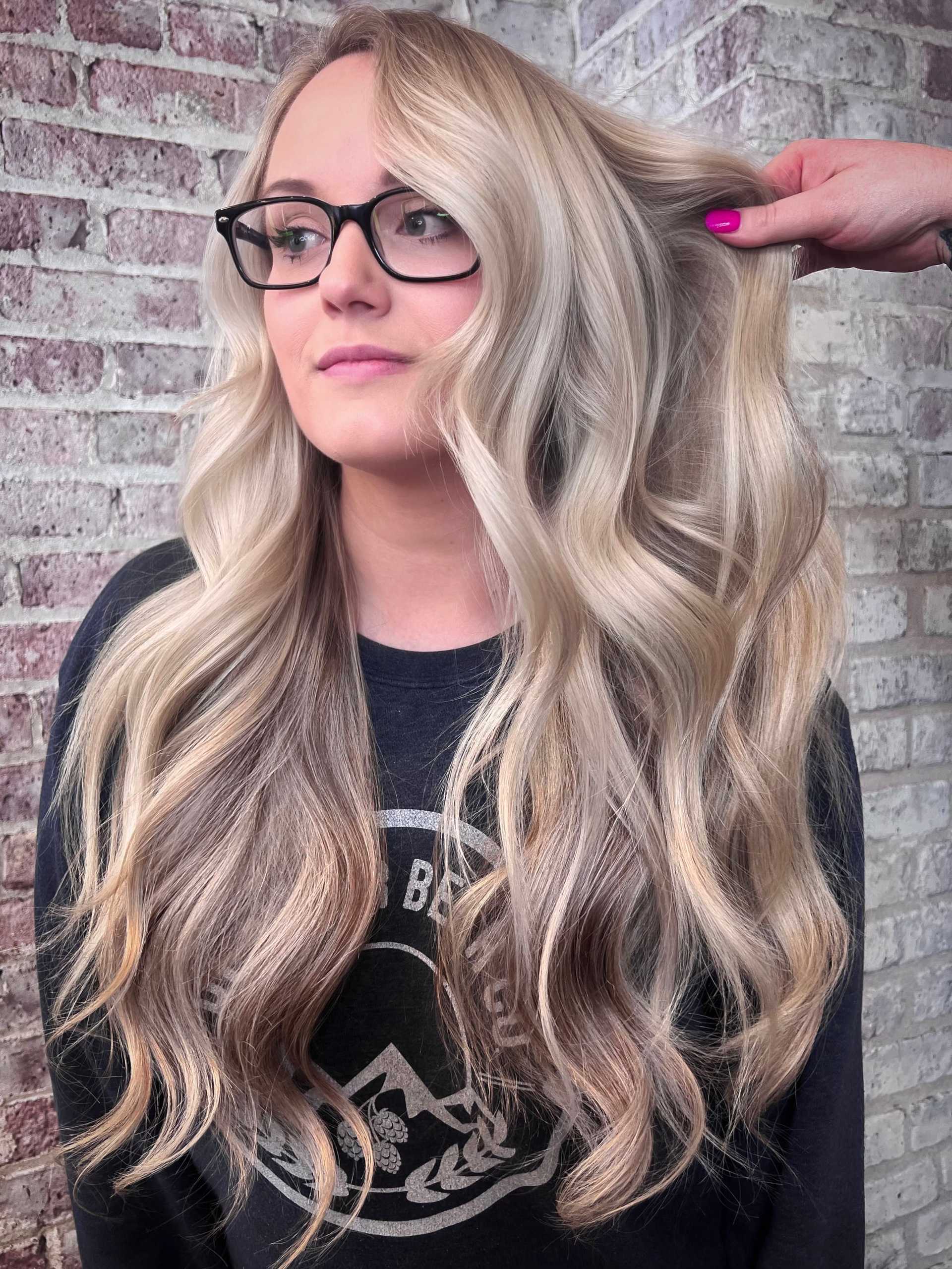 Woman with wavy blonde hair and black glasses in front of a brick wall.