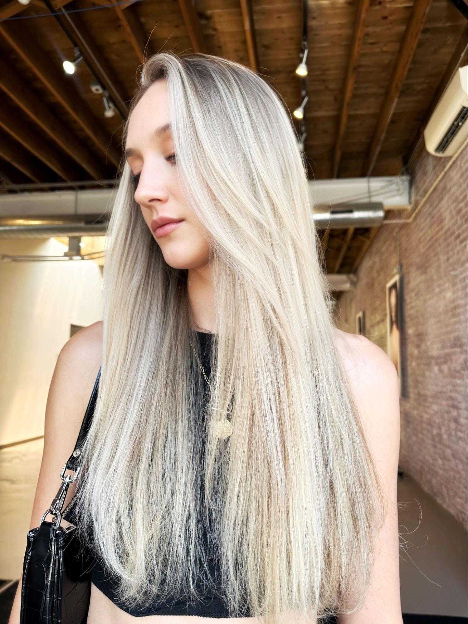 Young woman with long blonde hair in a brick-walled room, looking downward.
