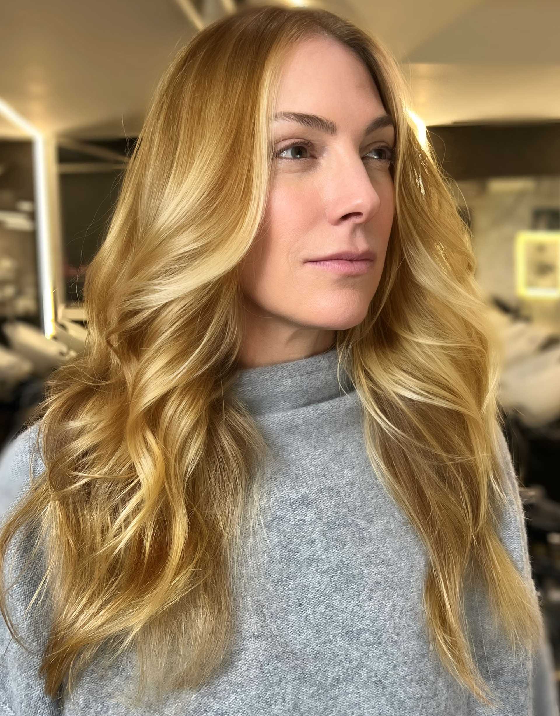 Blonde woman with wavy hair wearing a gray sweater in a salon setting.